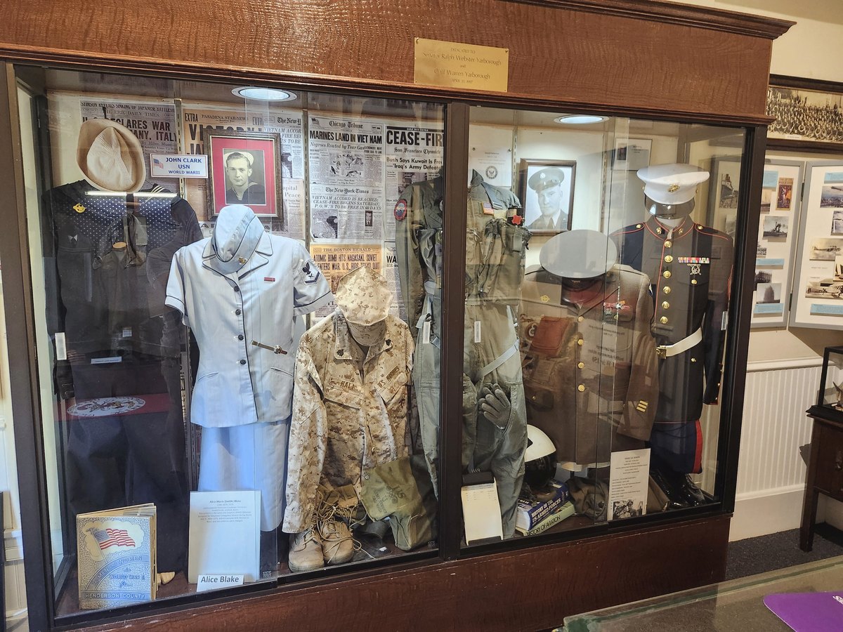 A long glass display case of donated military uniforms on mannequins, backed by old newspaper front pages and framed portraits