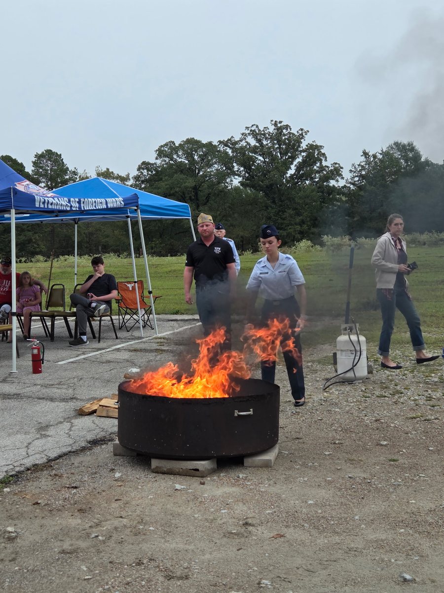 Flames rise from a round steel fire pit while a cadet in uniform and VFW Post 1799 Commander Paul Gray stand behind it, with a blue VFW tent and onlookers in the background