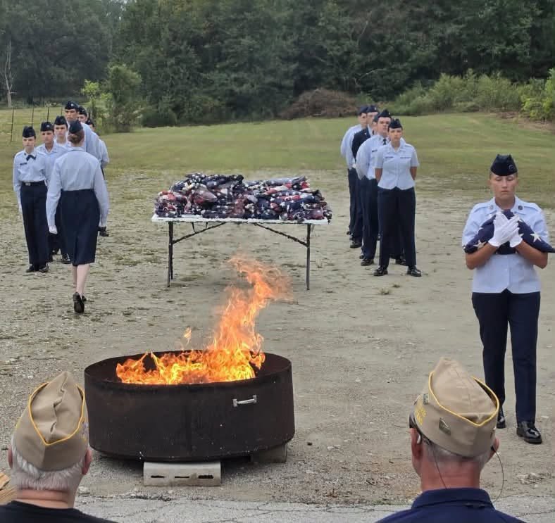 A cadet in white gloves holds a folded flag beside the burning fire pit, with a table of folded flags and cadets in formation behind and VFW members in caps in the foreground