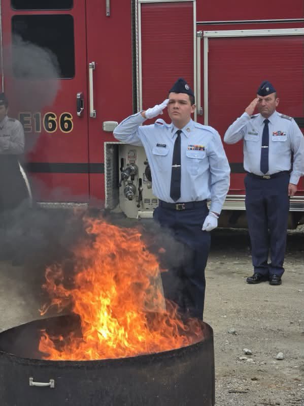 1st Lt. Trenton Klaus and a cadet salute beside the burning fire pit in front of a red fire engine marked E166