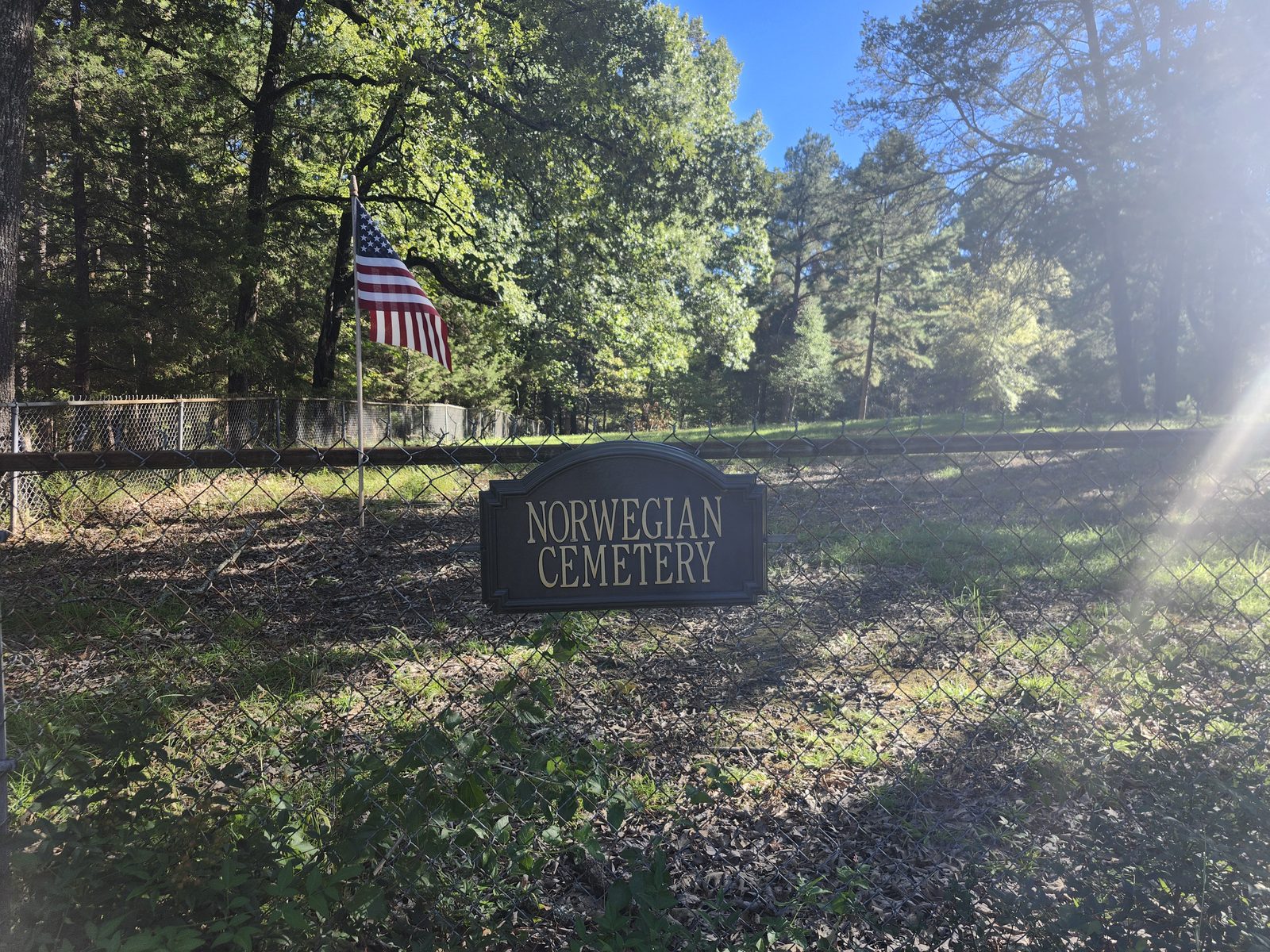 A “Norwegian Cemetery” sign on a chain-link fence near Brownsboro, with an American flag flying beyond